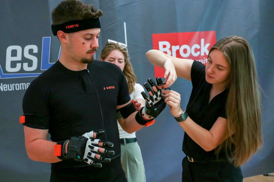 : A Brock University graduate student helps apply 3D motion capture equipment to her teammate’s hand. The part she is assisting with is a glove with sensors attached. 