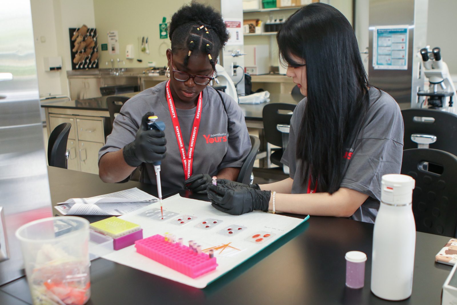 Two young women in a lab environment use a pipette to drop blood samples onto a dish for analysis.