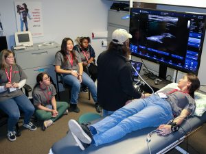 A man uses an ultrasound machine to show blood vessels on a large screen. A young woman lays on a table having the technique applied to her while four of her high-school aged peers look on.