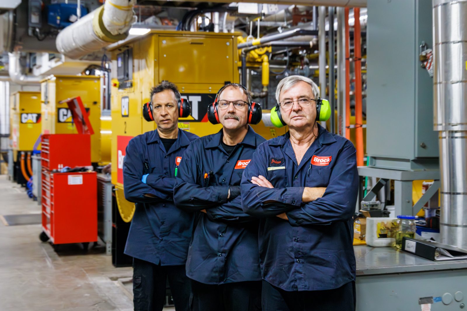 Three men stand in front of machinery in an engine room.