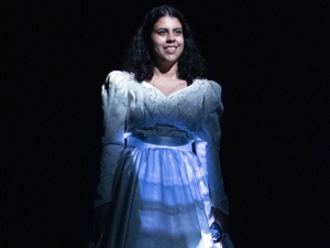 A woman in a white dress with the reflection of a video on it stands on a dark stage.