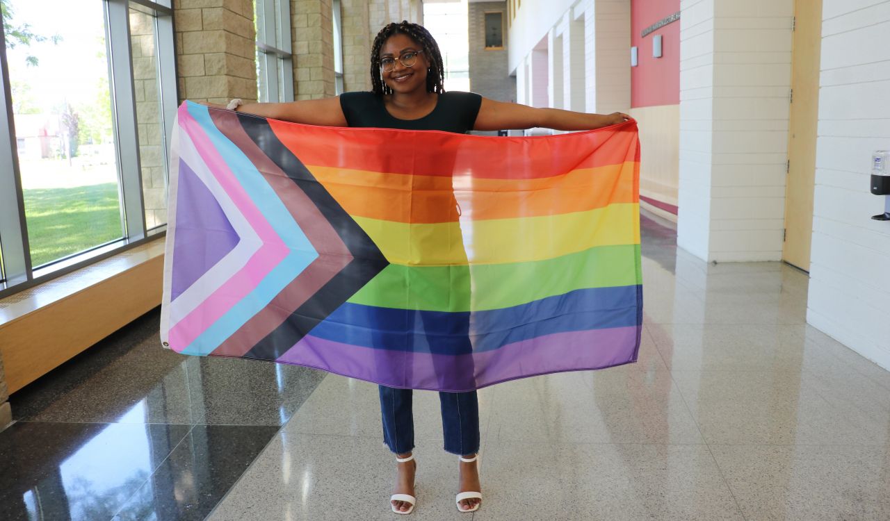 A woman stand in an indoor hallway holding a rainbow-coloured flag that also has multicoloured chevrons at one end.