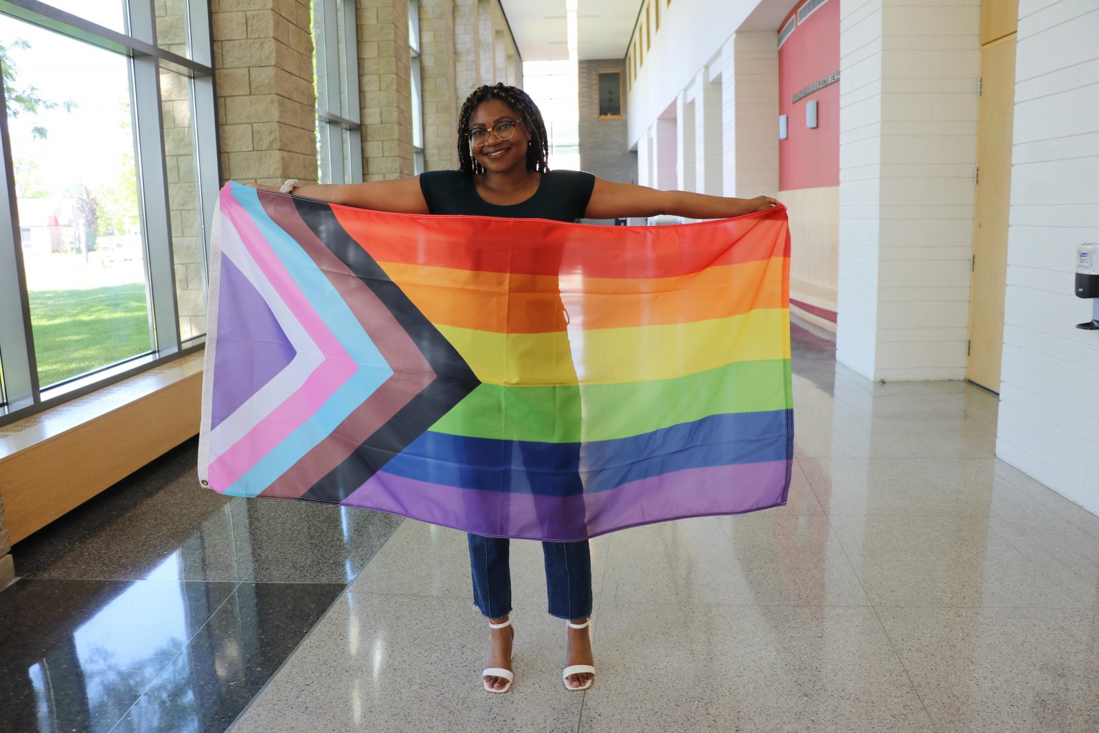 A woman stand in an indoor hallway holding a rainbow-coloured flag that also has multicoloured chevrons at one end.