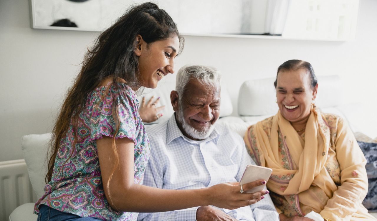 An elderly South Asian man wearing a striped vest looks down at a cellphone being held by a young female healthcare worker.