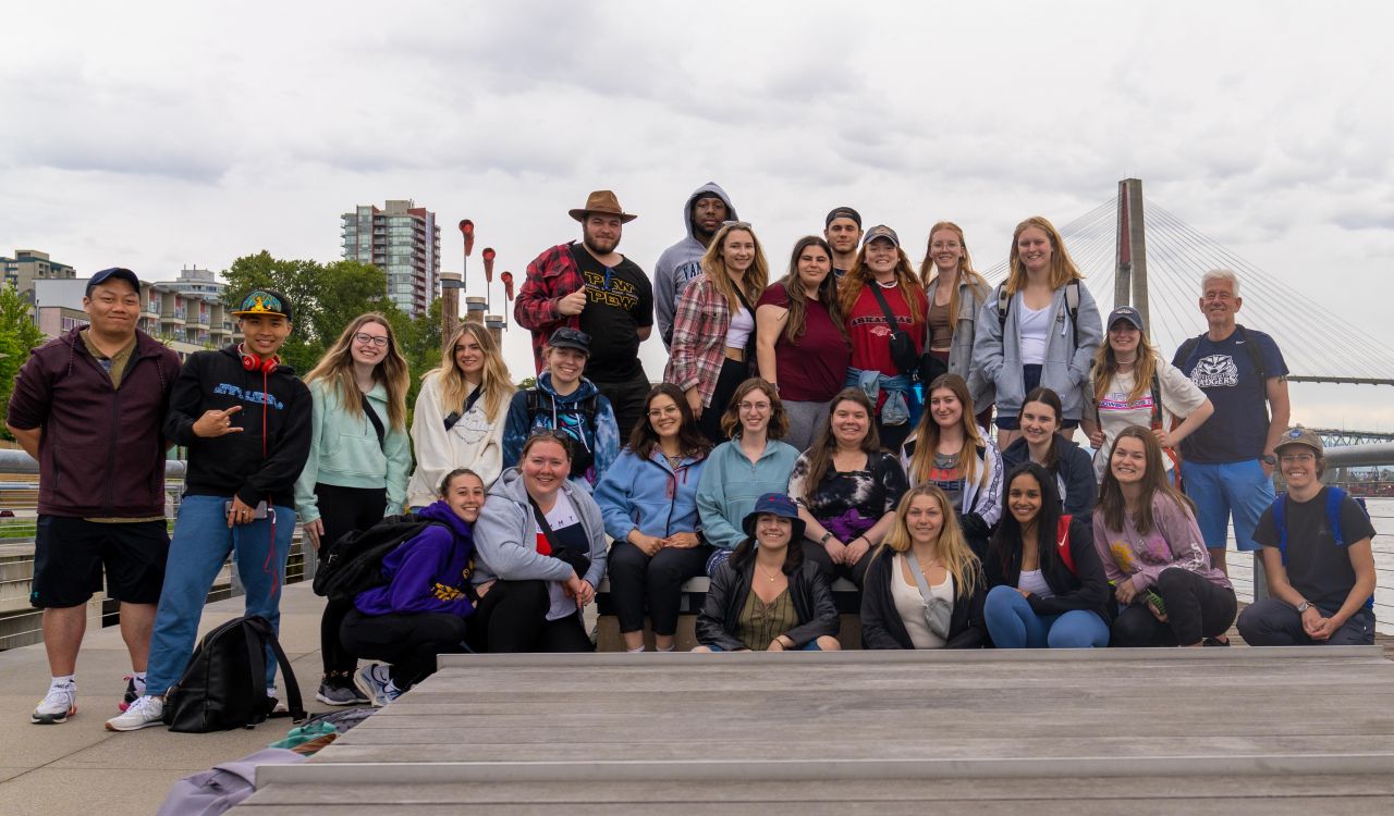 Students pose on boardwalk with Michael Ripmeester in front of a city skyline and bridge.