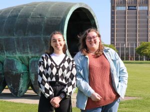 Two students stand beside a large metal sculpture with Brock University’s Schmon Tower in the background.
