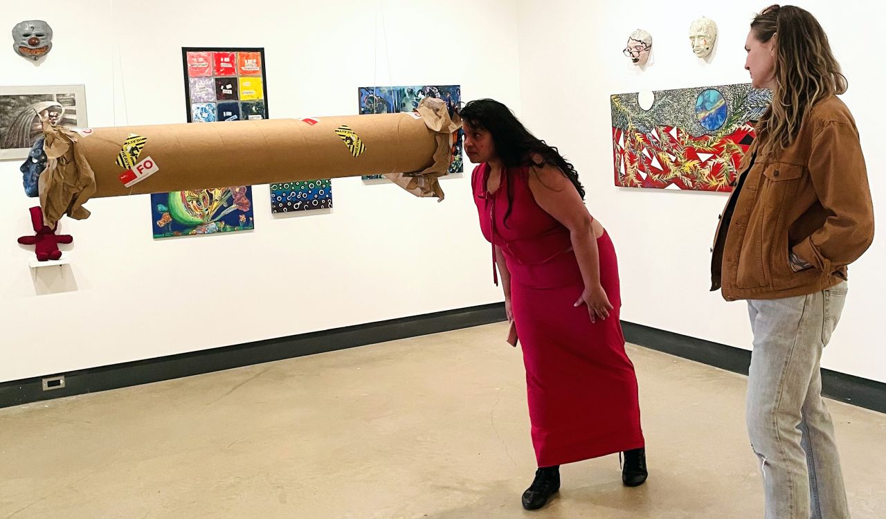 A woman in a red dress views a cylindrical piece of art hanging by strings from the ceiling in a bright, white art gallery while a second woman watches her. Colourful paintings hang on the walls around them.