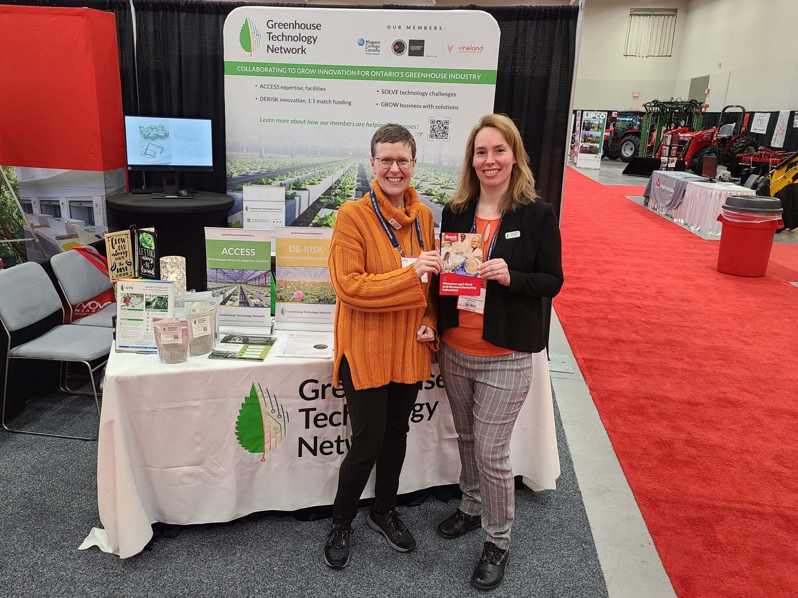 Two women stand in front of a conference display holding a brochure with a red carpet next to them and farming equipment in the background.
