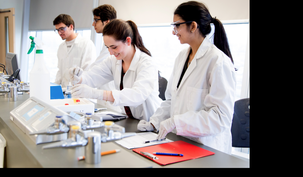 Four students wearing white lab coats work next to each other at a long table in a teaching laboratory.
