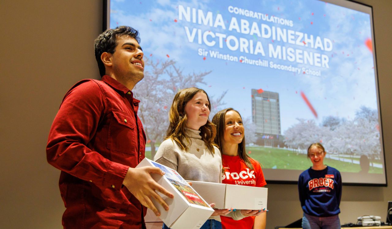 Three people stand together in front of a large projector screen that says congratulations. Two hold boxes in their hands.