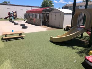 Sunny urban childcare centre playground with two areas of artificial grass with picnic tables and slides separated by an open play area with tires bordered by a shed and sheltering structure.