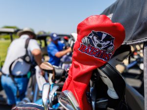 A close-up of a golf club with a red Brock Badgers cover. Players are picking up their bags in the background.