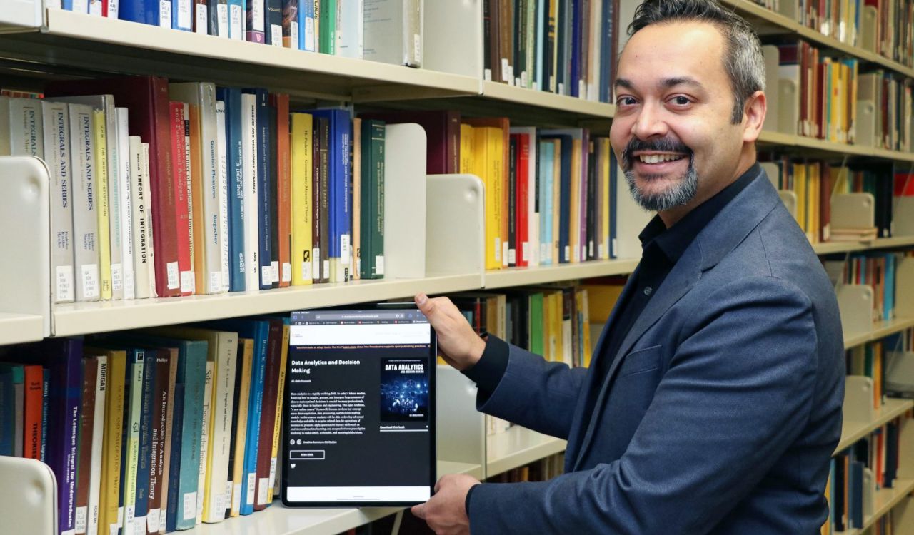 A man stands in a row of books in a library while holding a digital tablet.