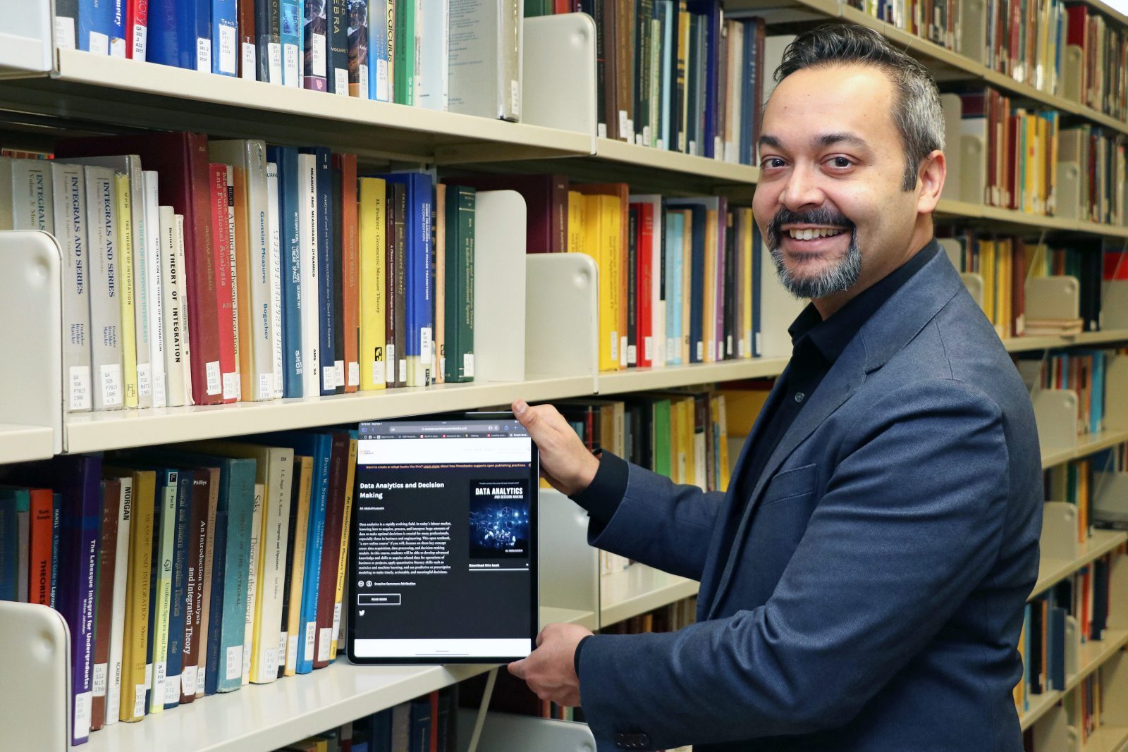 A man stands in a row of books in a library while holding a digital tablet.