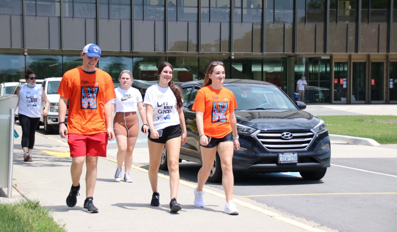 A group of people in white and orange shirts walks along a sidewalk at Brock University.