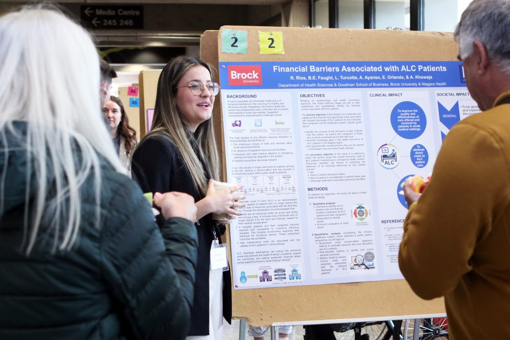 A young women stands next to a large poster displaying a lot of text about heath-care research as she speaks to two people.