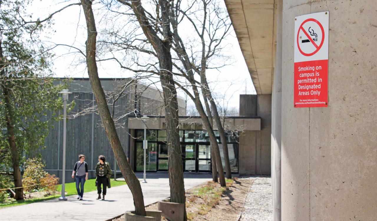 A sign on the exterior of a building reads "Smoking on campus is permitted in designated areas only." Two people are visible walking outside in the background.