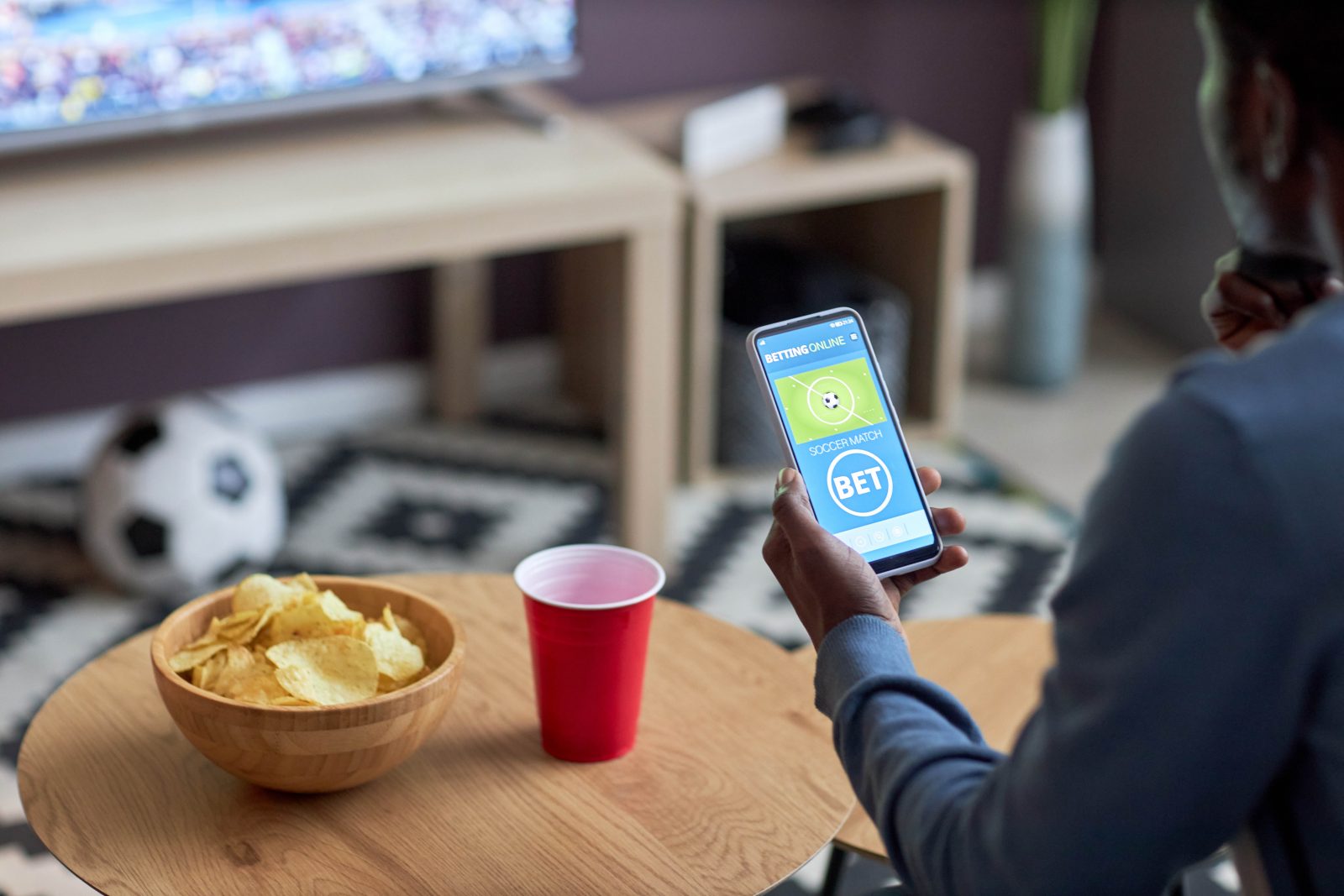 A hand holds a phone displaying that a bet has been placed next to food on a table with a blurred television in the background.