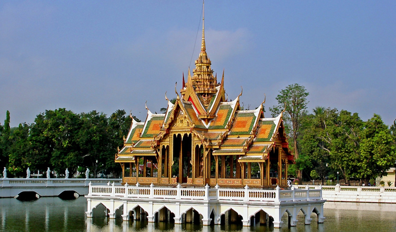 Bang Pa-In Royal Palace elevated on a white platform in the Chao Phraya River in Thailand, with trees and a blue sky in the background.