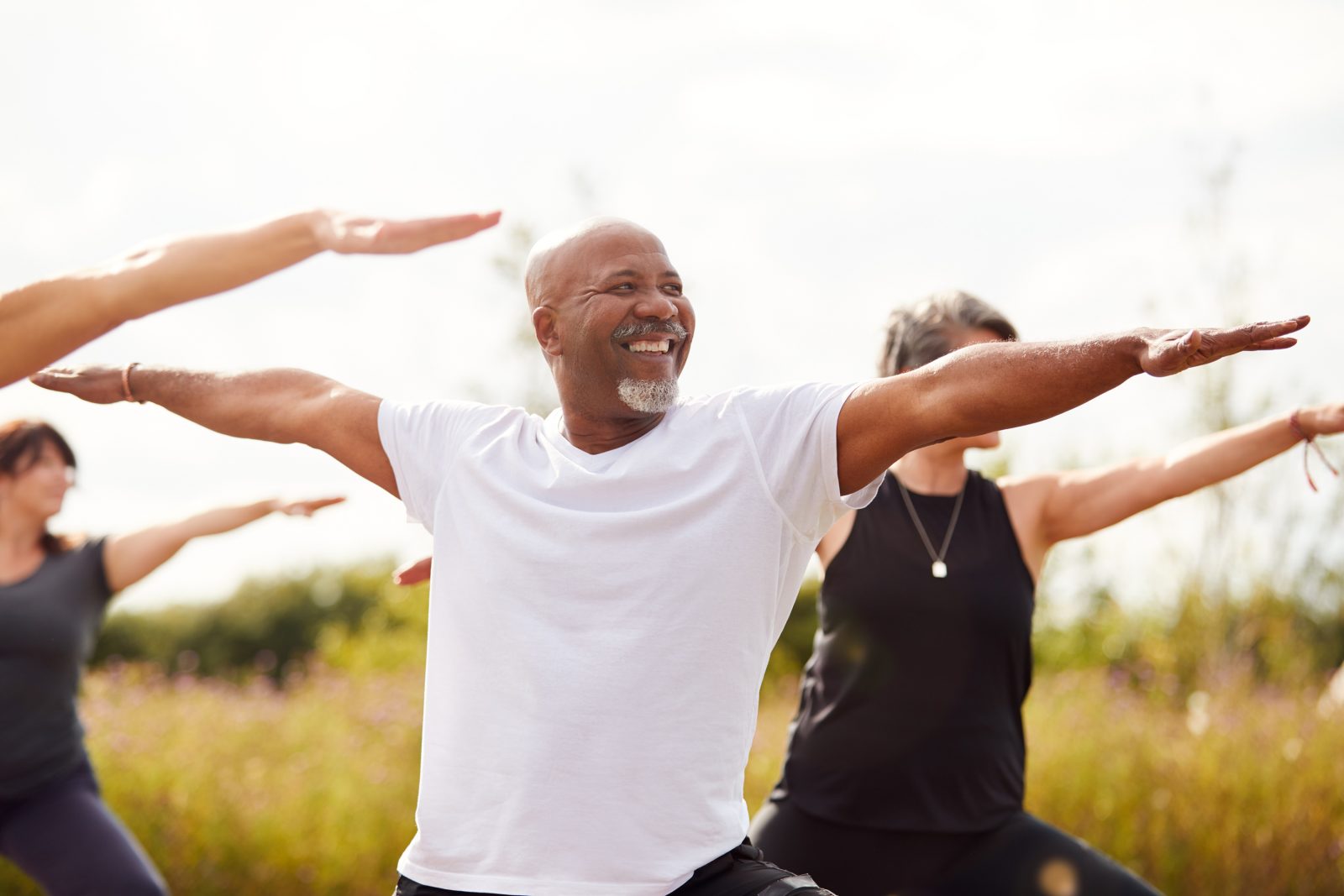 An older man does yoga outside with a group of other people of varying ages and genders. The man has a smile on his face as he performs warrior 2 pose, with his arms stretched straight out from his shoulders, parallel to the ground, and his face looking towards one of his hands.