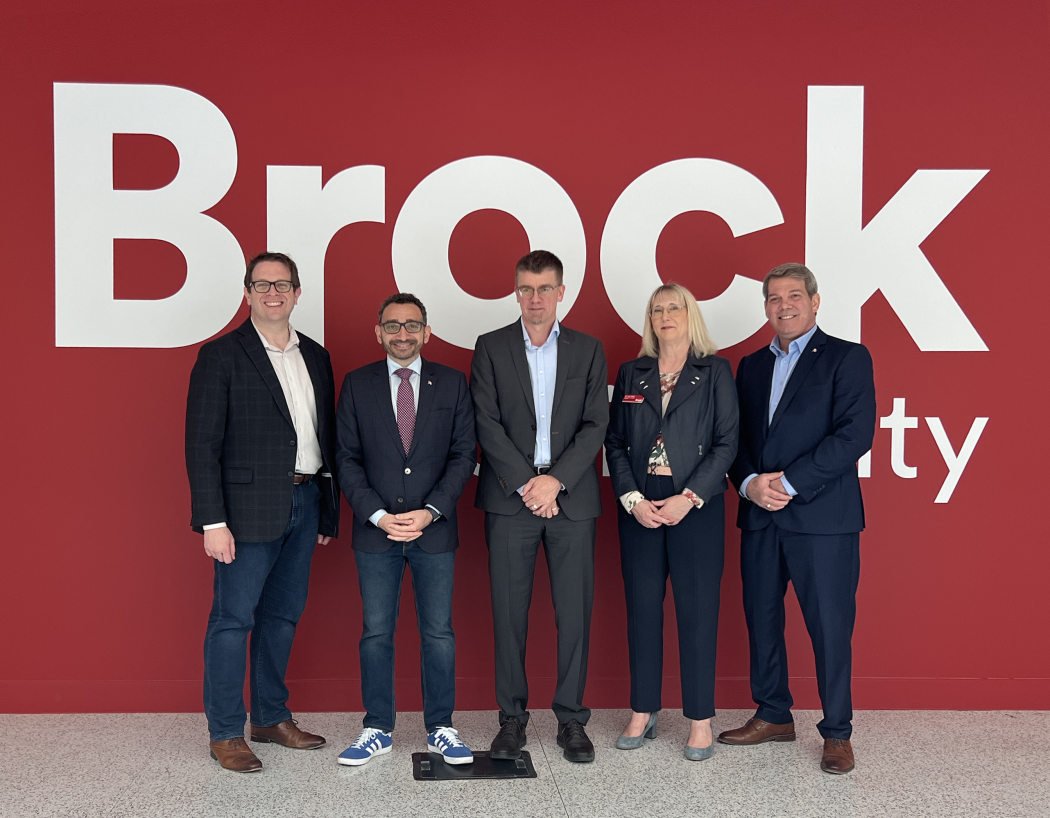Five people stand side-by-side in front of a large red wall with the white Brock University logo on it.