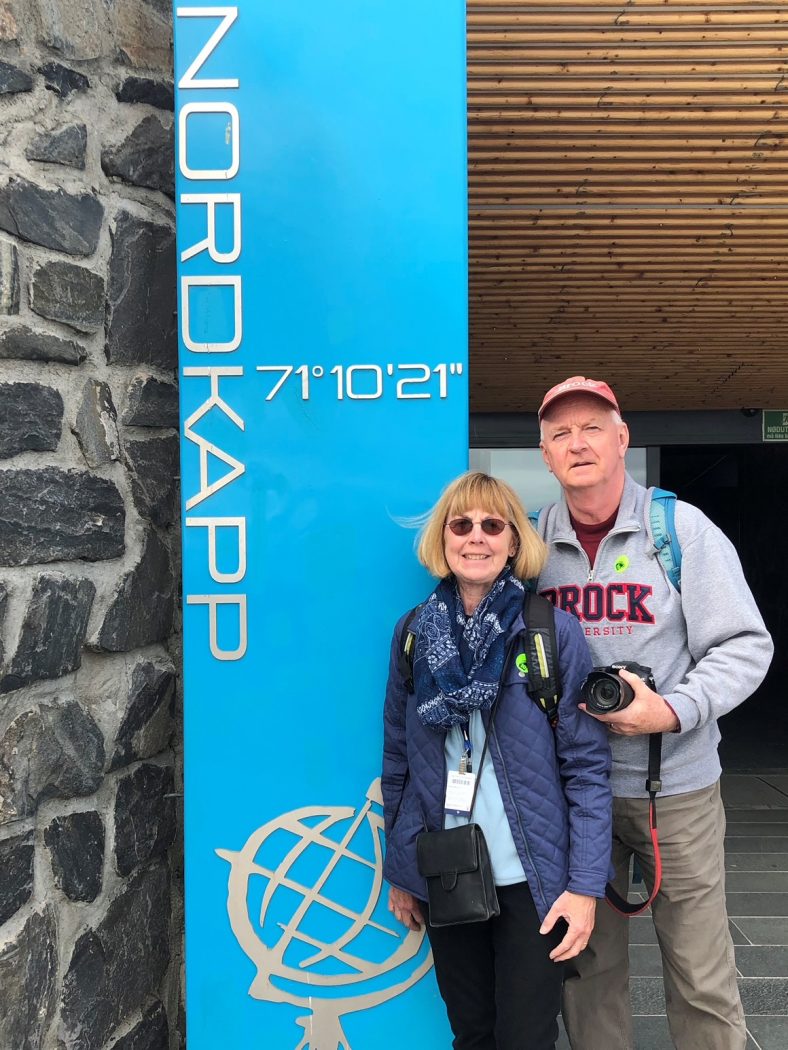 Trish and Greg Finn stand in front of a sign that says “NORDKAPP 70°10’21”” in Norway. 