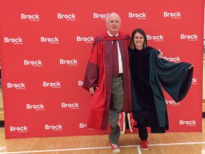 Standing on one foot in front of a Brock branded backdrop and wearing academic robes, Greg Finn and Geraldine Jones touch together the heel of one shoe to the heel of the other person.