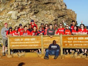 A group of 24 people stand at the base of a mountain with the ocean behind them and a large wooden sign that says “Cape of Good Hope,” which is the most south-western point of the African Continent.