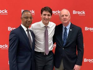 Three men dressed in business attire stand in front of sign with Brock University logos on it.