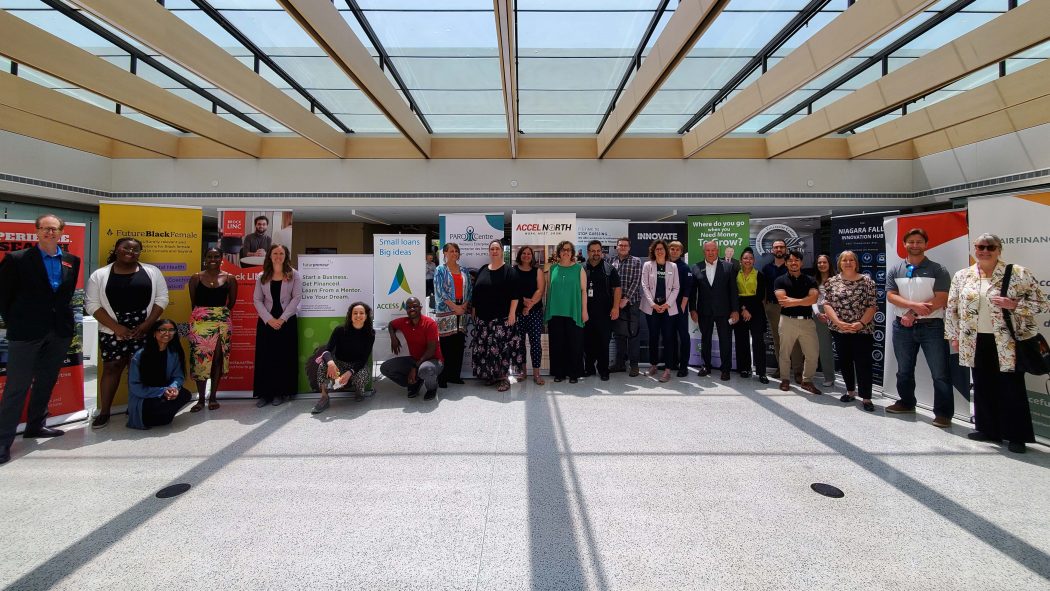 A group of entrepreneurs stand beside each other in a sunny atrium. Promotional banners are visible behind them.
