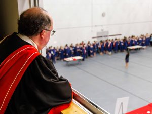 A man in academic regalia looks out into a gym full of university students wearing graduation robes.