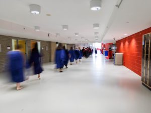 A line of people wearing graduation robes walk into a university convocation ceremony.