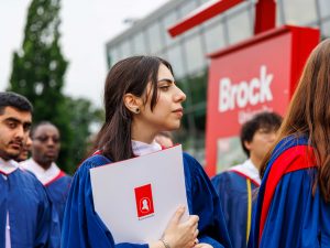 A woman wearing graduation robes holds her diploma at Brock University's Convocation. Her fellow graduates mingle nearby.