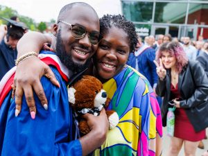 A new graduate in academic robs holds a graduation bear and hugs his loved one. A friend smiles at the camera behind the two people.