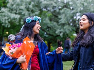 A new graduate in Convocation robes holds a bouquet of flowers and smiles at a loved one.