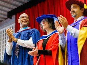 Three people in academic regalia applaud on stage during a graduation ceremony.