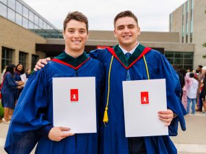 Two Brock graduates pose in their gowns while holding their diplomas.