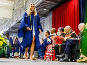 A smiling student in a graduation gown walks across the stage at Convocation while faculty and guests applaud.