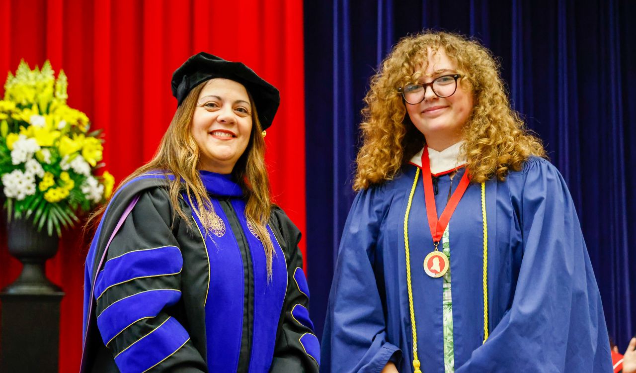 A woman in academic regalia stands beside young woman wearing a graduation gown and a medal.