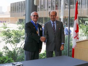 Two men stand in front of bright windows shaking hands with a table in front of them with pins. The man on left wears a medal.