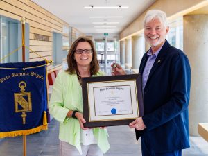A blue sign with the words Beta Gamma Sigma hangs to the left of Brock President and Vice-Chancellor Lesley Rigg’s shoulder as she is presented a framed award and pin from Goodman Dean Barry Wright, who is wearing the same key-shaped pin.