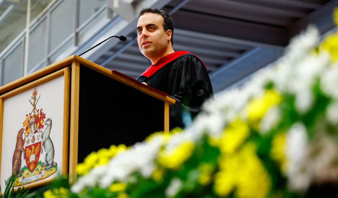 Barak Shoshany, dressed in academic regalia, speaking at a podium on a stage during a Brock University Convocation ceremony.