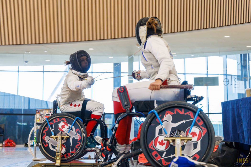 Two fencers in wheelchairs compete with swords while surrounded by sunlit windows.