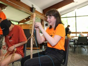 A woman in an orange shirt weaves leather and string into a traditional wooden lacrosse stick, while sitting next to another woman who is doing the same thing.
