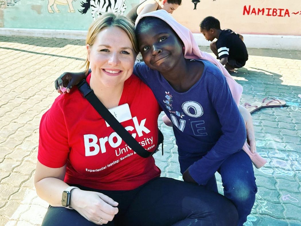 A woman wearing a red Brock University T-shirt and a child crouch down on a sidewalk that has been decorated with chalk. They smile for a photo as they embrace each other, each with one arm around the other’s shoulders. 