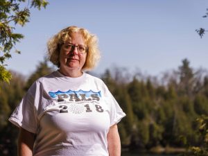 A woman in a white T-shirt and glasses stands next to a lake with trees in the background.