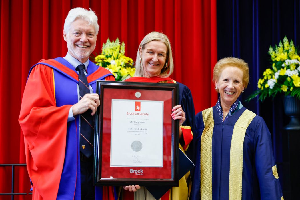 Three people in Convocation gowns stand holding a framed award.
