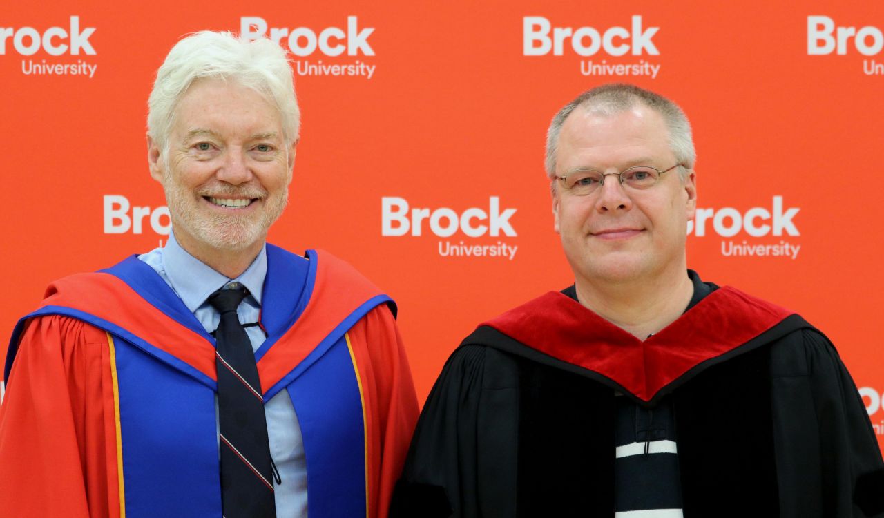 Dean of the Goodman School of Business Barry Wright and Professor of Management Dirk De Clercq stand in front of a Brock University banner. Both men are wearing academic regalia.