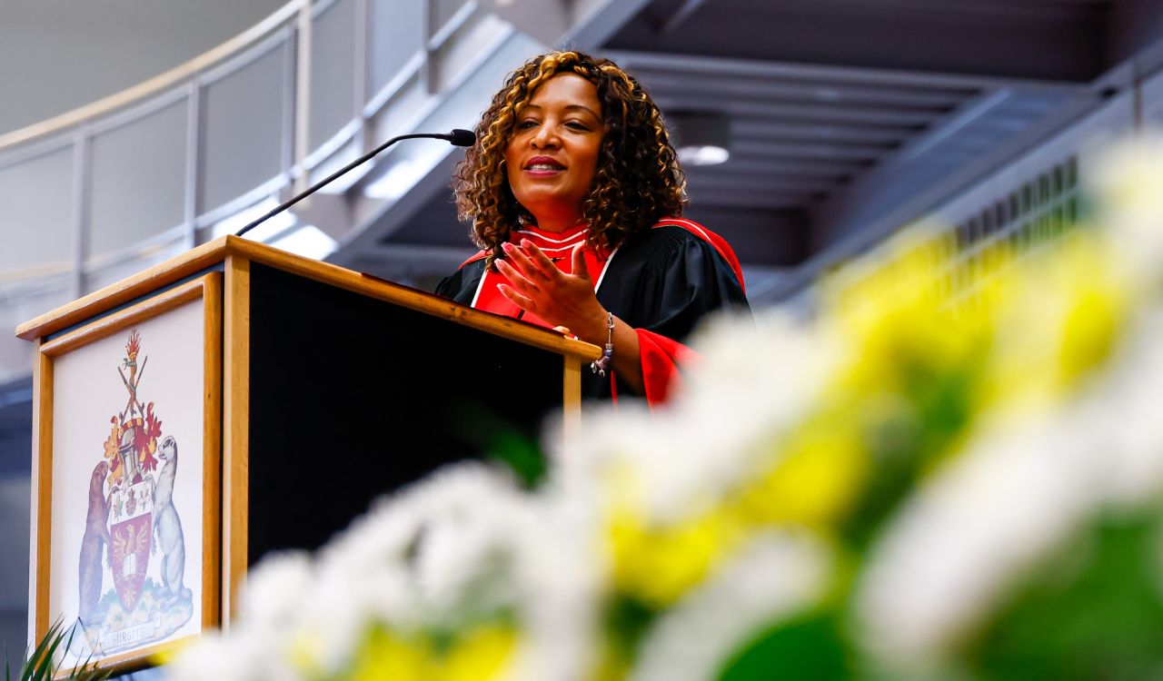 A woman in academic regalia speaks from behind a podium during a graduation ceremony.