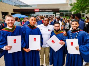 A group of four young men in graduation gowns pose for a photo with a friend and their diplomas after Convocation.